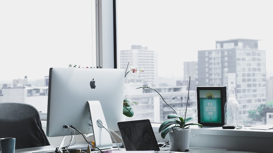 sunlit desk with an iMac and a small plant beside a window overlooking the city